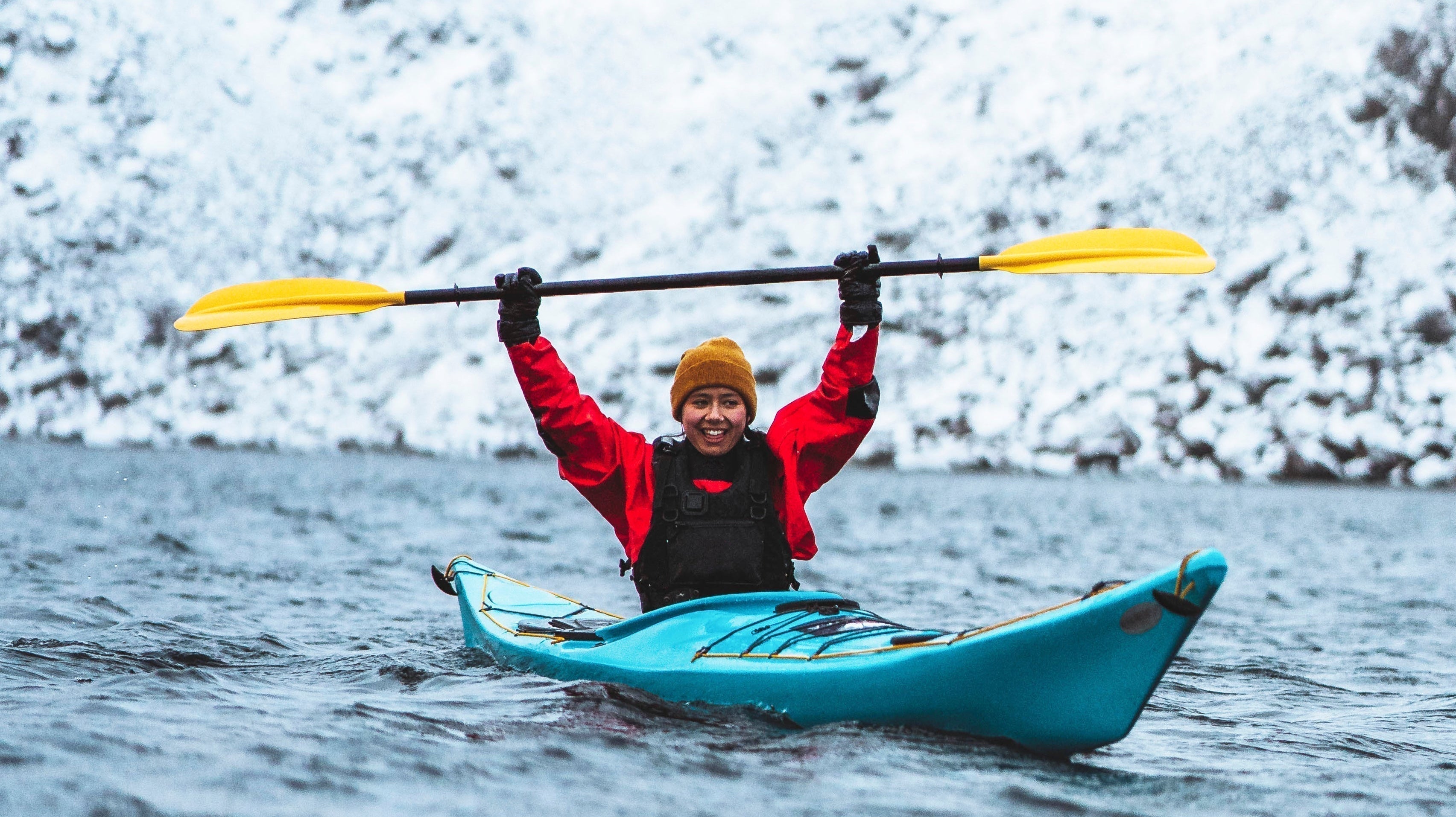 Woman kayaking in cold water wearing a red drysuit and yellow beanie, snowy fjord background
